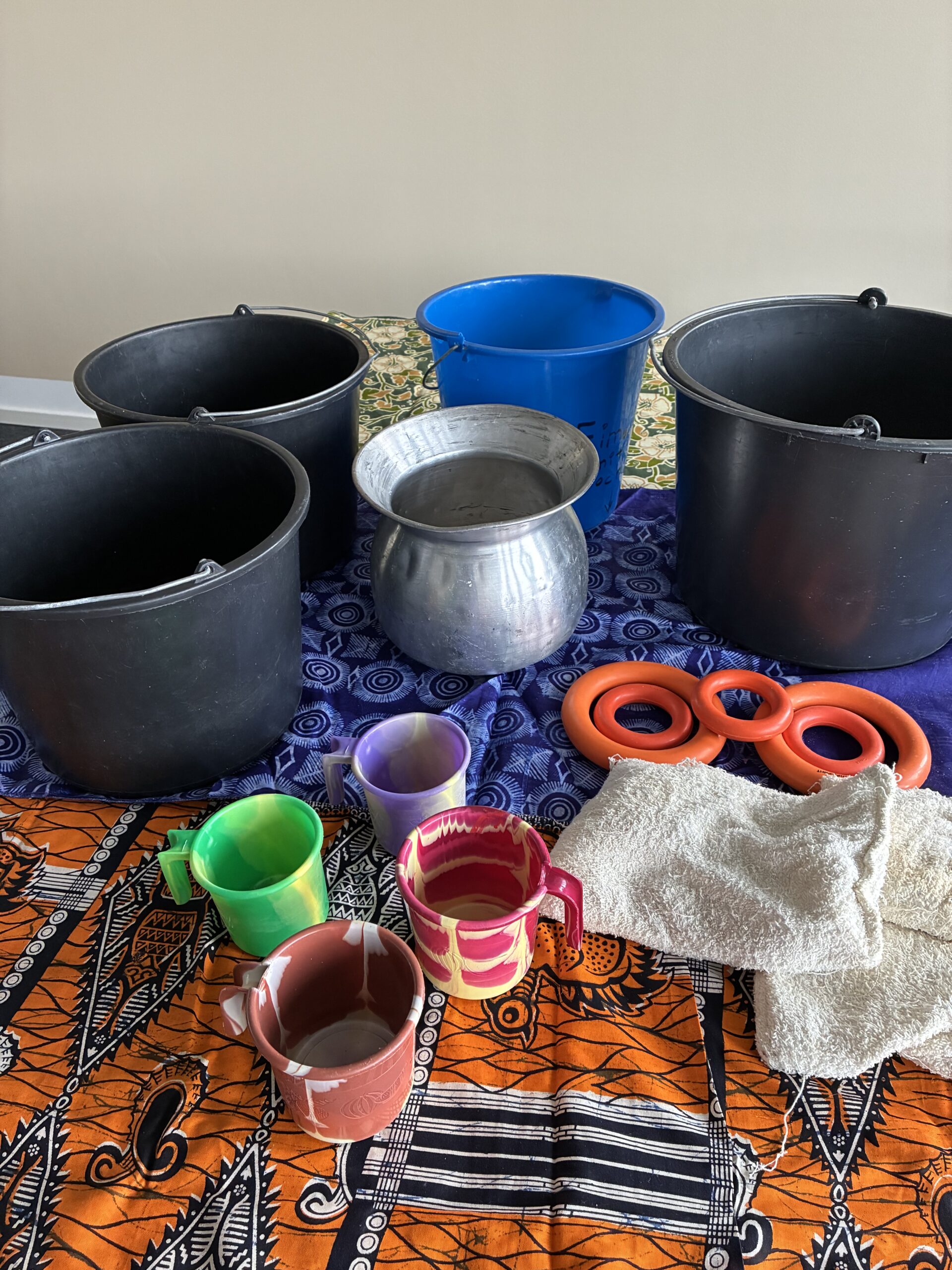Das Aktionsset „Wasserholen Afrika“ umfasst Plastikeimer, einen Metalltopf, bunte Becher, orangefarbene Deckel und ein weißes Handtuch, die auf einem orange-blau gemusterten Tuch vor einer beigen Wand angeordnet sind.