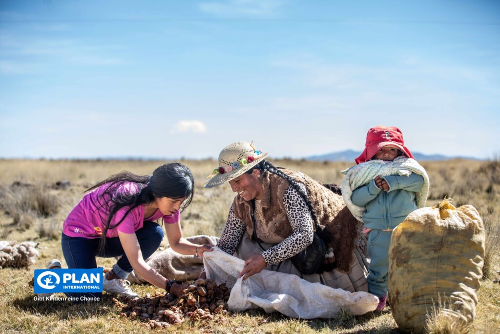 A woman and a young girl sort potatoes in a field, smiling together. A small child stands nearby, wrapped in warm clothes. Large sacks are on the ground, and the sky is clear. A Plan International logo appears in the corner.