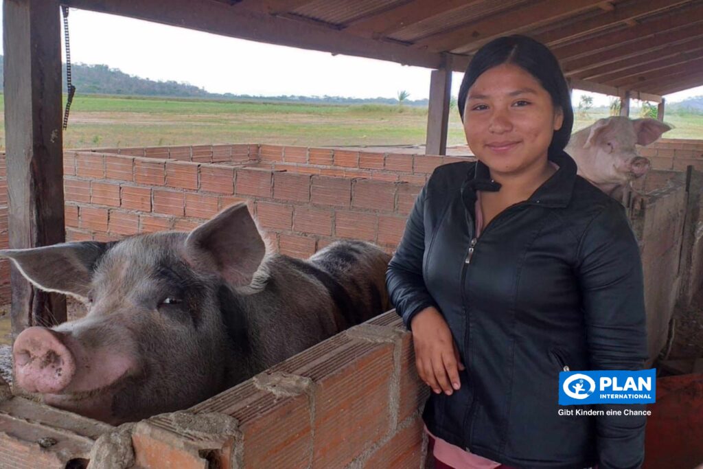 A young woman stands smiling next to a pig pen with two large pigs inside. She is wearing a black jacket and is under a wooden roof in a rural setting. The Plan International logo appears in the lower right corner.