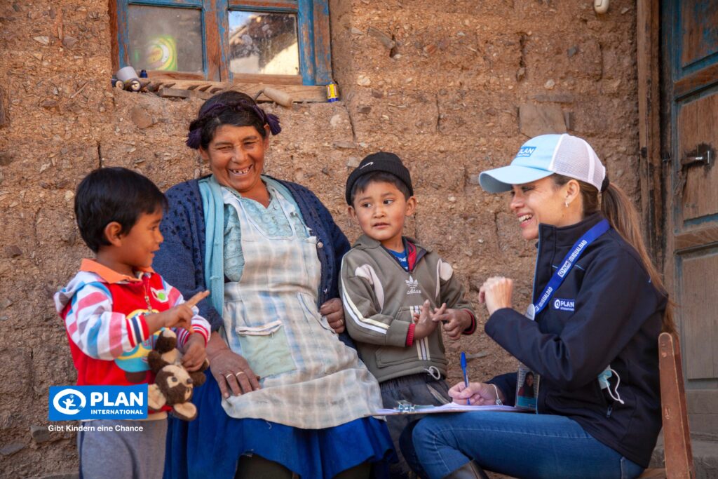 A woman and two children smile and interact with a woman in a Plan International cap and jacket, who is writing on a clipboard outside a rustic building. The children appear happy and playful.