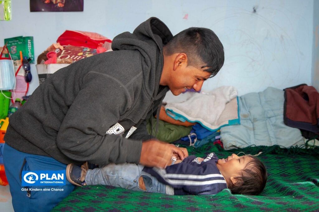 A smiling man plays with a laughing toddler lying on a green blanket in a colorful room. The logo of Plan International is visible in the lower left corner.