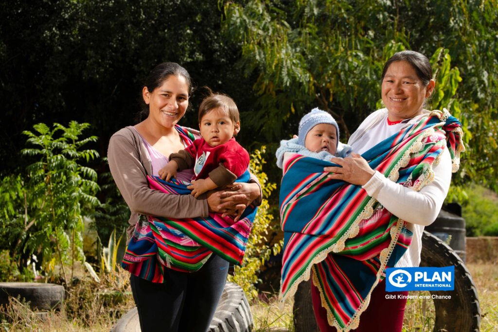 Two women stand outdoors, each holding a baby in a colorful, striped traditional cloth. They are smiling, with green trees in the background. The Plan International logo appears in the lower right corner.
