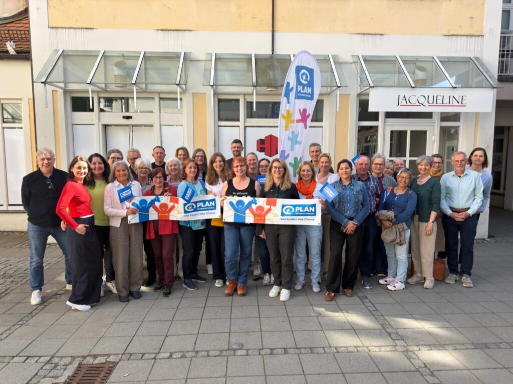 Eine große Gruppe von Menschen steht lächelnd im Freien vor einem Gebäude mit einem Schild mit der Aufschrift JACQUELINE. Einige halten PLAN-Banner und Schilder mit blauen und orangefarbenen Handabdrücken. Im Hintergrund ist eine große Flagge von Plan International zu sehen.