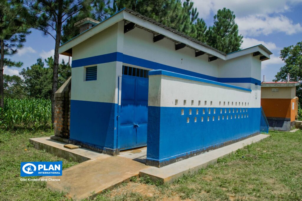 Ein blau-weißes Gebäude mit einer geschlossenen blauen Tür steht auf einer Wiese in der Nähe von Bäumen und Maisfeldern. In der unteren linken Ecke ist das Logo von Plan International zu sehen.