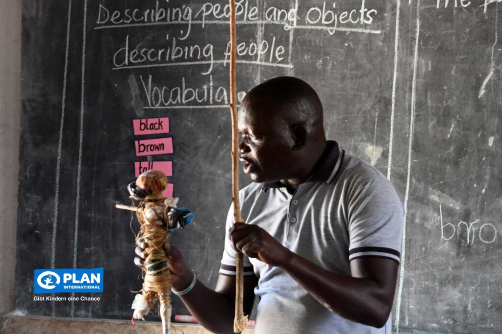 Eine Lehrerin, die eine selbstgemachte Puppe in der Hand hält, gestikuliert, während sie vor einer Tafel mit englischen Vokabeln unterrichtet. In der unteren linken Ecke ist das Logo von Plan International zu sehen.
