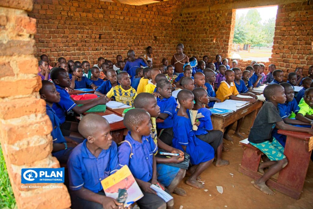 Eine große Gruppe von Schulkindern in blauen und gelben Uniformen sitzt in einem Klassenzimmer aus Backstein mit offenen Fenstern, hört aufmerksam zu und hält Bücher in den Händen, während ein Lehrer hinten im Raum steht. In der Ecke ist das Logo von Plan International zu sehen.