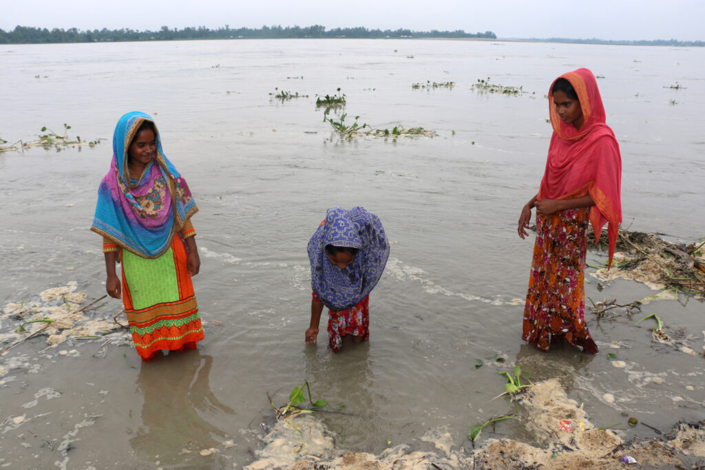 Drei Frauen in farbenfroher Kleidung stehen am Rand eines überfluteten Gebiets. Eine beugt sich vor und berührt das Wasser, während die anderen beiden zusehen. Im Hintergrund sind die weite Wasserfläche und ein bewölkter Himmel zu sehen.
